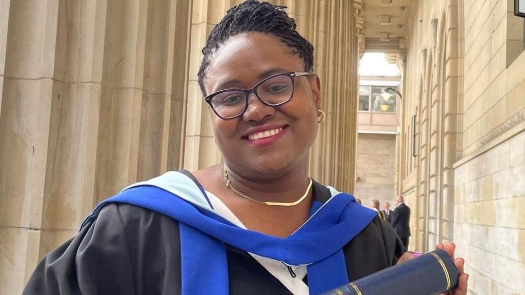 Dr Gomo, who is wearing black graduation robes, smiles as she holds a University of Dundee scroll. She is also wearing glasses.