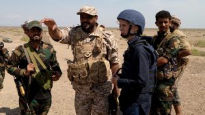 Commander Tawfiq Al Haboun in combat fatigues gestures next to Orla Guerin wearing blue in a group of armed people wearing combat fatigues