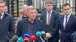 A man with short dark grey hair speaking to the media wearing a black blazer and polo shirt.