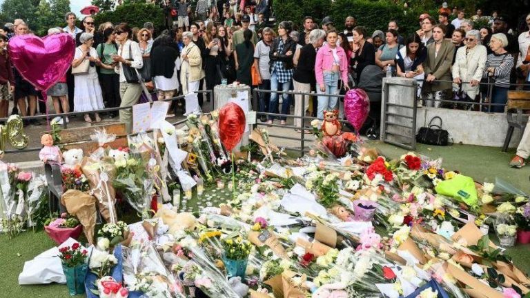 A large crowd of people gather behind a patch of grass in the Annecy park which is covered with bouquets of flowers, balloons and teddy bears.