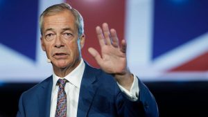 Nigel Farage holds his hand up while speaking on a stage with a Union flag in the background.