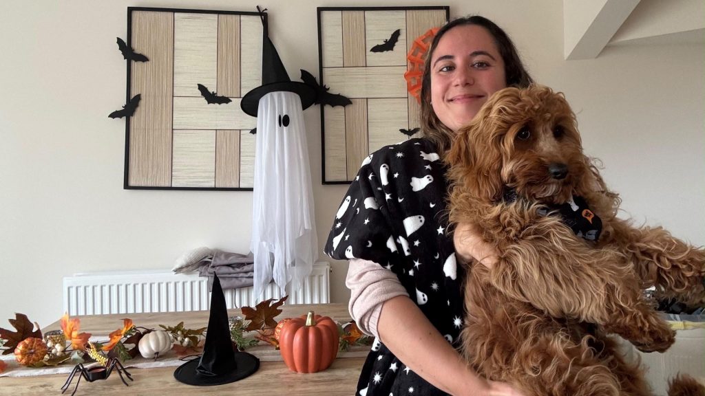 A young woman shows off her spooky Halloween decorations in her living room as she holds her puppy and smiles. She is wearing a black scarf with drawings of white ghosts. Behind her, a table with a witch's hat and pumpkin decorations, a fake spider, and hung on the wall is a ghost wearing a witch's hat surrounded by bats.