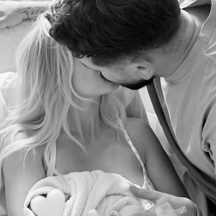 A black and white image showing a man with dark hair kissing his wife with blonde hair. The woman is cradling a baby wrapped in a towel in her arms.
