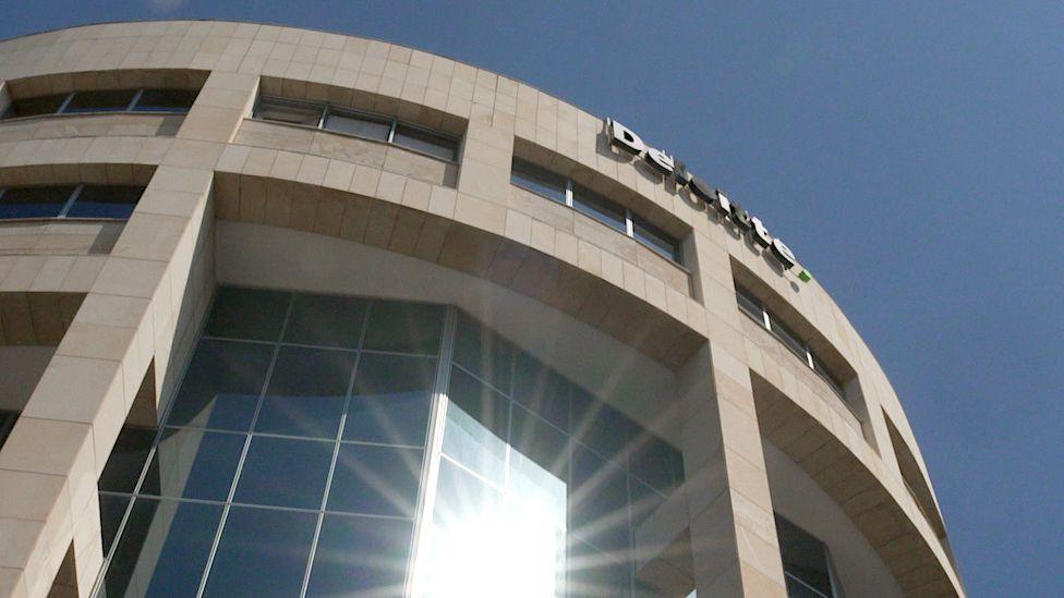 A view of Deloitte's offices in Limassol, Cyprus, looking up at a tall, curved building in honey-coloured stone and tinted glass, with blue sky above and a sunburst of reflected light at the bottom of the image.