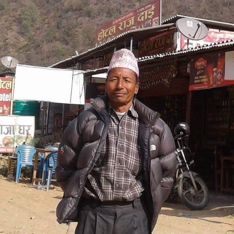 A man in a black puffer jacket, chequered shirt and hat stands outside in a street in front of buildings