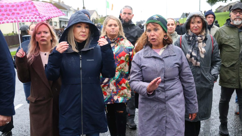 A crowd of people walk down the Garvaghy Road, at the front is a woman wearing a navy coat and another woman wearing a grey coat