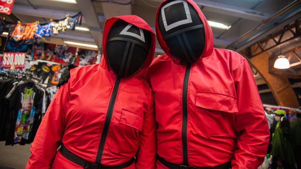 Two people wear red boiler suits and black masks from Squid Game inside a costume shop