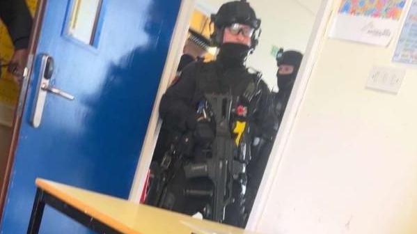 An armed police officer is at a doorway to a classroom. He is wearing all black clothing with a helmet and glasses. He is holding a large black gun, pointed towards the floor