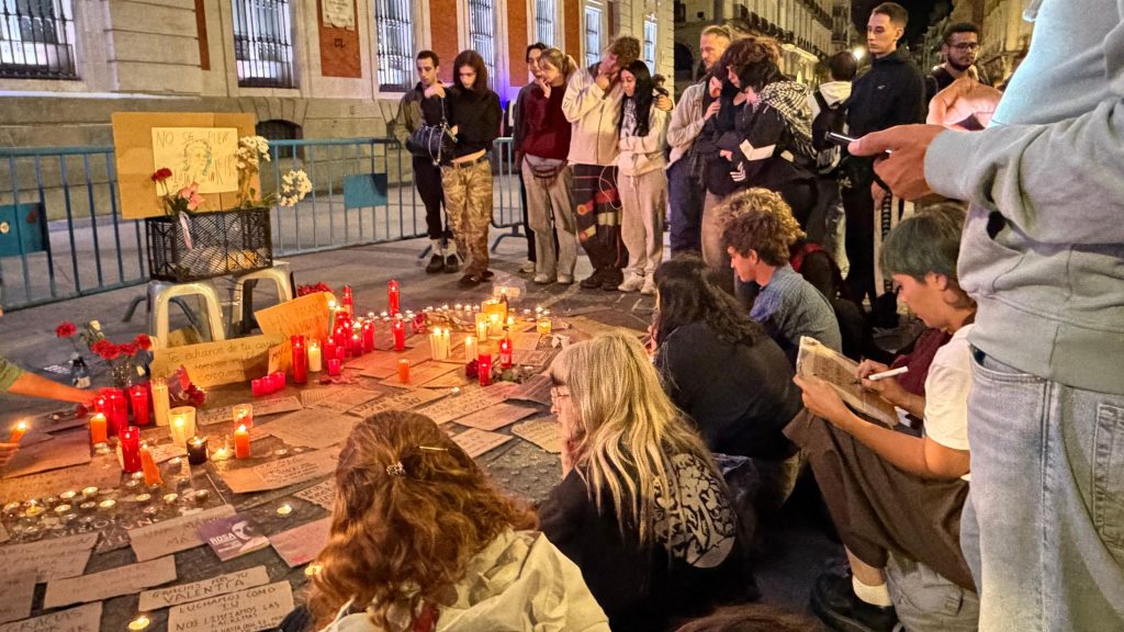 People gather around a vigil where red candles and written tributes sit on pavement near a fence.