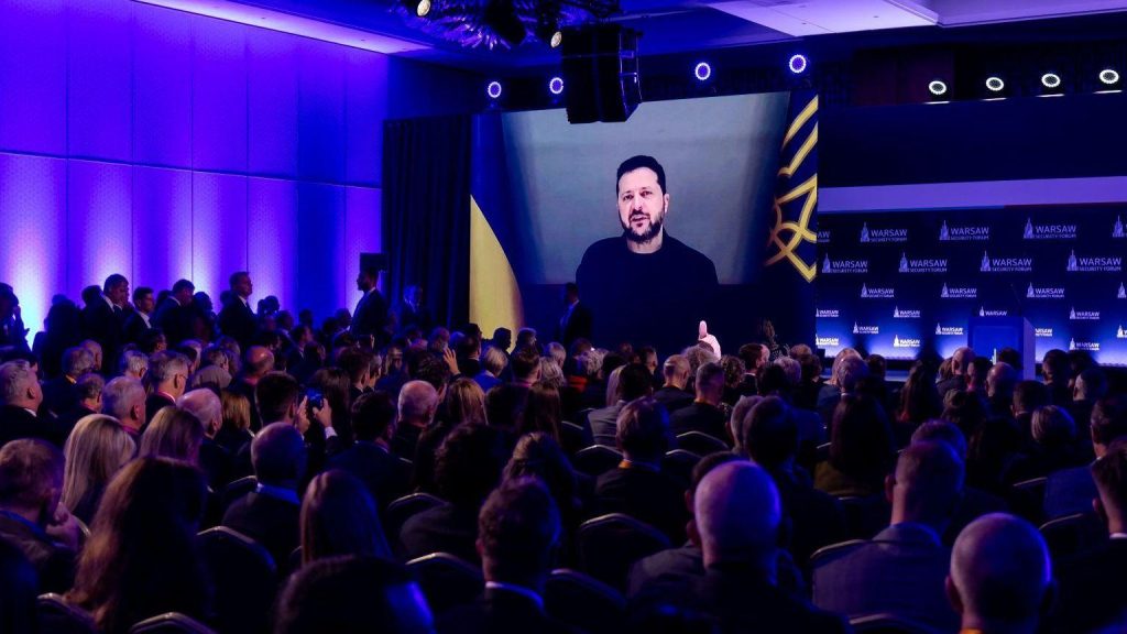 Volodymyr Zelensky speaking via video on a projector in front of audience members at the Warsaw Security Forum. Zelensky is wearing black while about 100 people in the audience watch him on the screen with blue lighting in the room