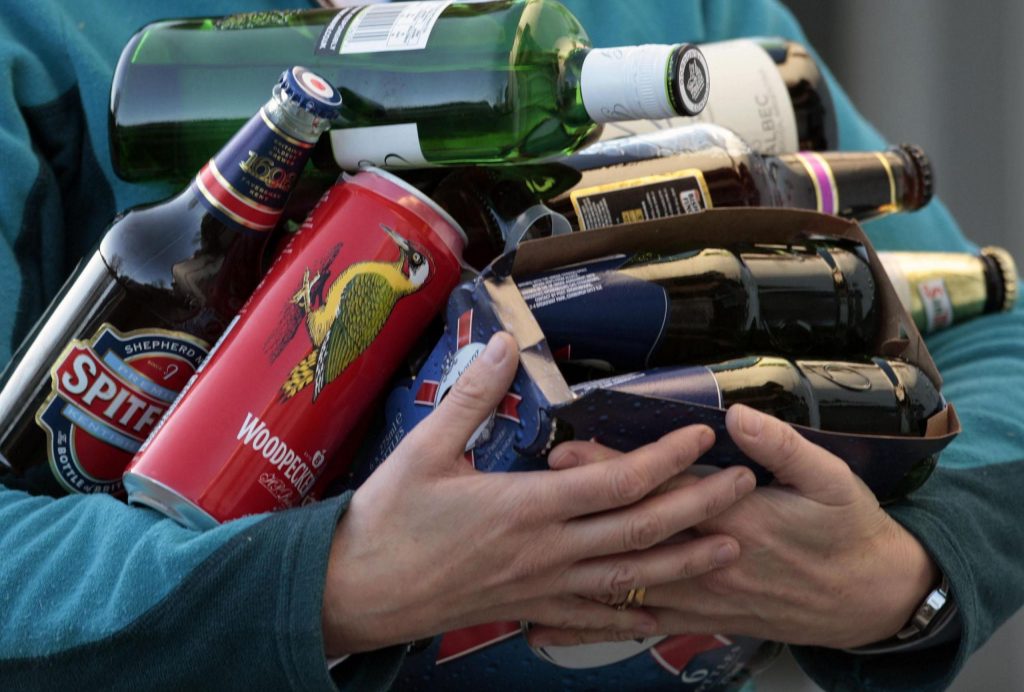 A picture of a man struggling to carry several types of alcohol bottles - including various beers as well as red wine and gin. The man, whose face we cannot see, is wearing a light blue jumper.