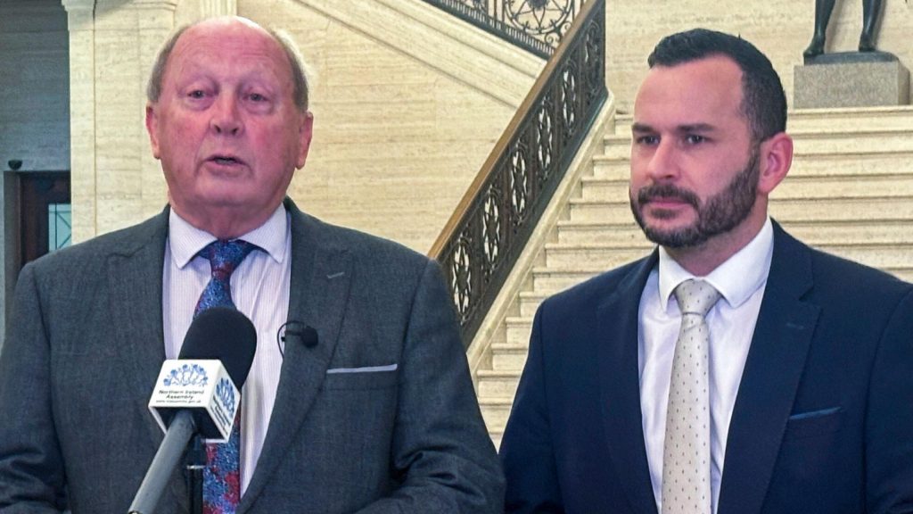 Two men standing at a microphone, the man on the left has short hair and is wearing a grey suit with blue and red tie and the man on the right has dark hair and beard, he is wearing a navy suit and gey tie with dots