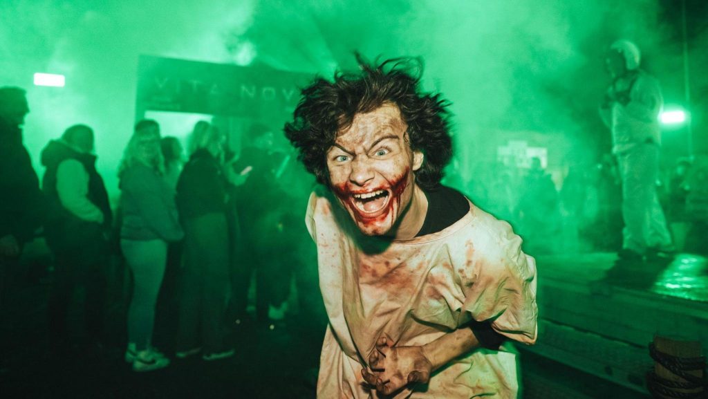 A scary-looking actor has blood all over his face and wears a bloody, loose straight jacket. He looks like he just came out an asylum. Behind him there's a crowd of people and lots of green smoke.