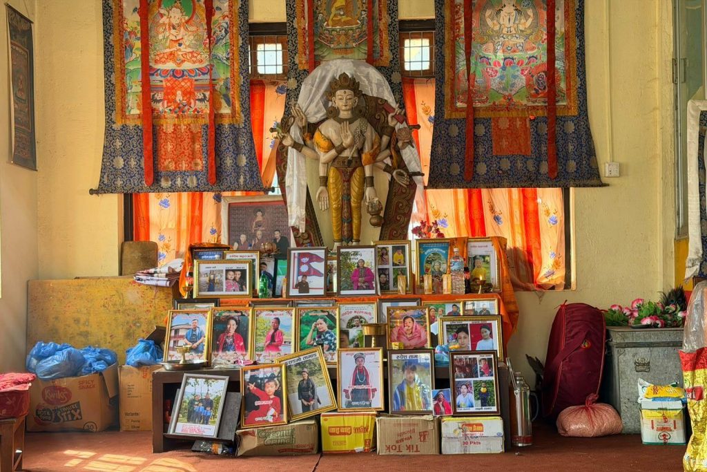 Dozens of framed photographs arranged beneath the figure of a religious deity inside a building, with multicoloured and patterned curtains hanging in the background