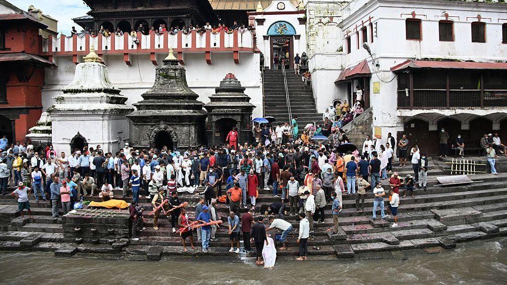 A crowd gathers along the stone steps of a riverside cremation site beside several small temple shrines and white buildings. People stand and watch from the steps, balconies and surrounding walkways, while the river flows in the foreground