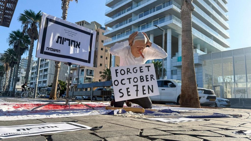 A protester wearing a face mask depicting Israeli Prime Minister Benjamin Netanyahu kneels on a sheet covered with red paint with a sign in Hebrew announcing the "untimely death" of "responsibility", in Tel Aviv, Israel (7 October 2026)