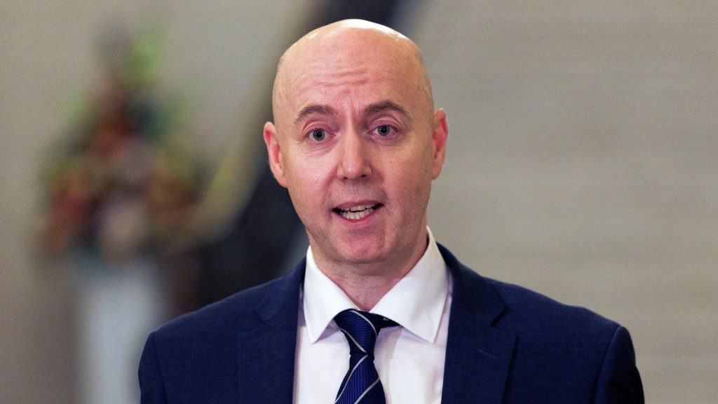 Jon Burrows is wearing a white shirt with navy blazer and tie. The background is blurry.